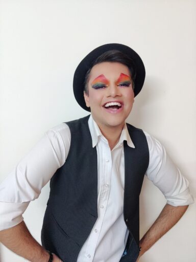 Aryan in a white shirt with a black vest standing with their hands on hips smiling with a black hat on and amazing rainbow eye shadow.